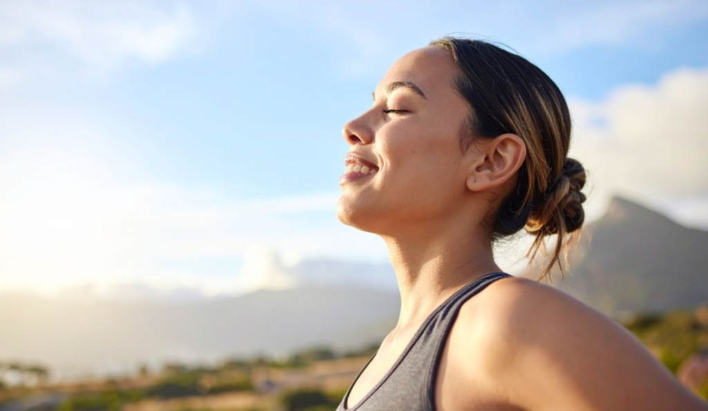 Smiling woman standing in sunlight with eyes closed, embracing warmth and practicing acceptance and letting go of control