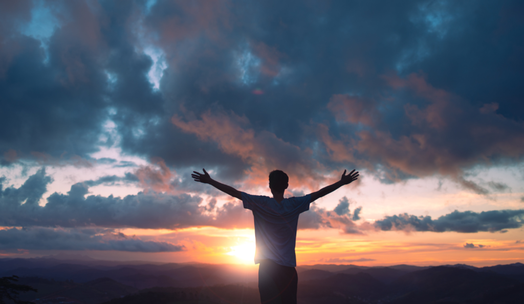 Man standing on a mountain at sunrise with arms outstretched, symbolizing freedom, openness, and gratitude