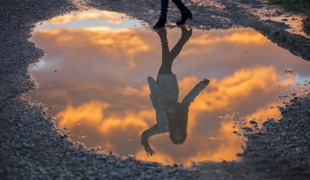 Woman standing beside a puddle with her reflection waving back, symbolizing self-distancing, self-awareness, and perspective shift
