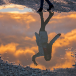 Woman standing beside a puddle with her reflection waving back, symbolizing self-distancing, self-awareness, and perspective shift