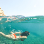 Woman swimming underwater beside a rocky jetty, surrounded by ocean currents and sunlight filtering through the water.