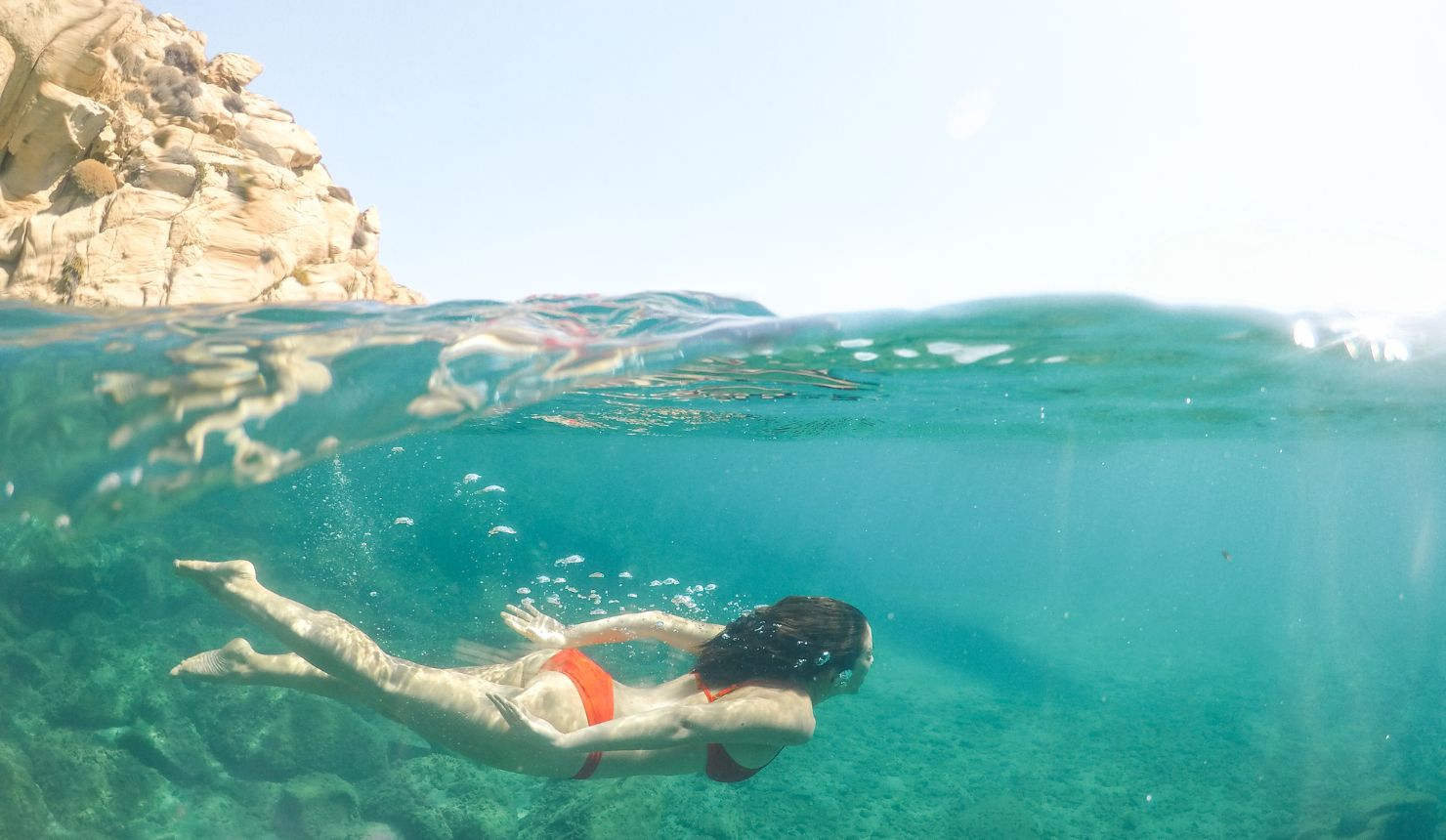 Woman swimming underwater beside a rocky jetty, surrounded by ocean currents and sunlight filtering through the water.