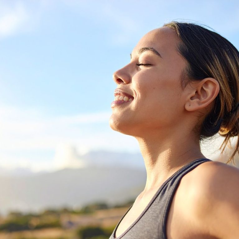 Smiling woman standing in sunlight with eyes closed, embracing warmth and practicing acceptance and letting go of control