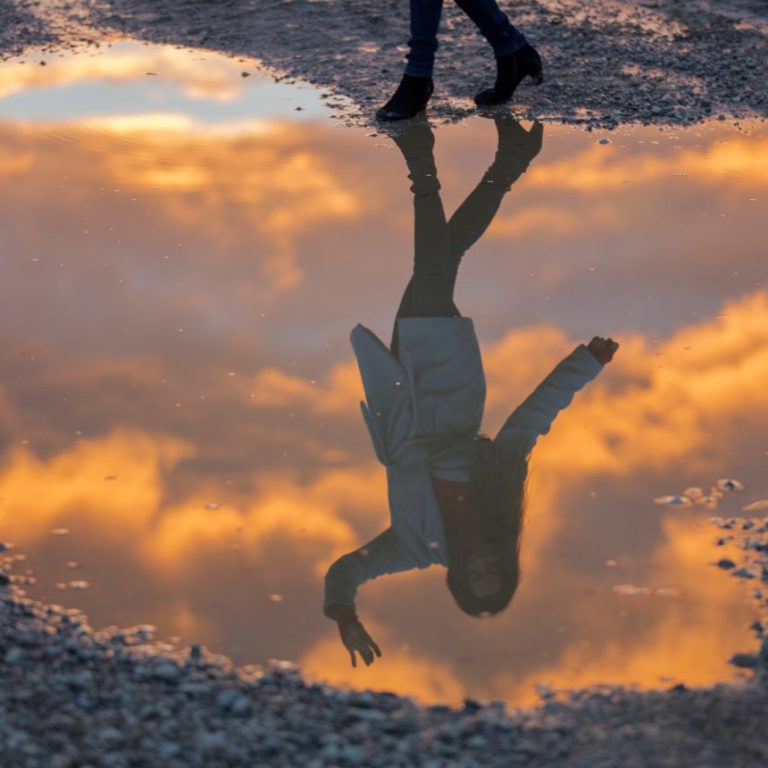Woman standing beside a puddle with her reflection waving back, symbolizing self-distancing, self-awareness, and perspective shift