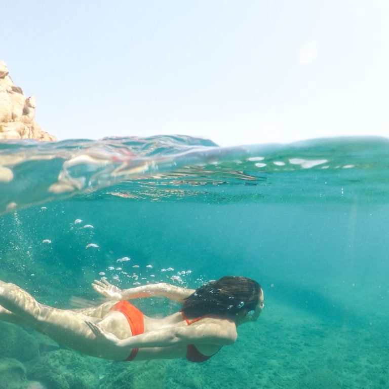 Woman swimming underwater beside a rocky jetty, surrounded by ocean currents and sunlight filtering through the water.